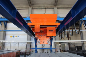  The orange-colored concrete distributor is suspended from the two blue steel supports of the portal for traveling over the entire length of the mold situated below and filling it with concrete  